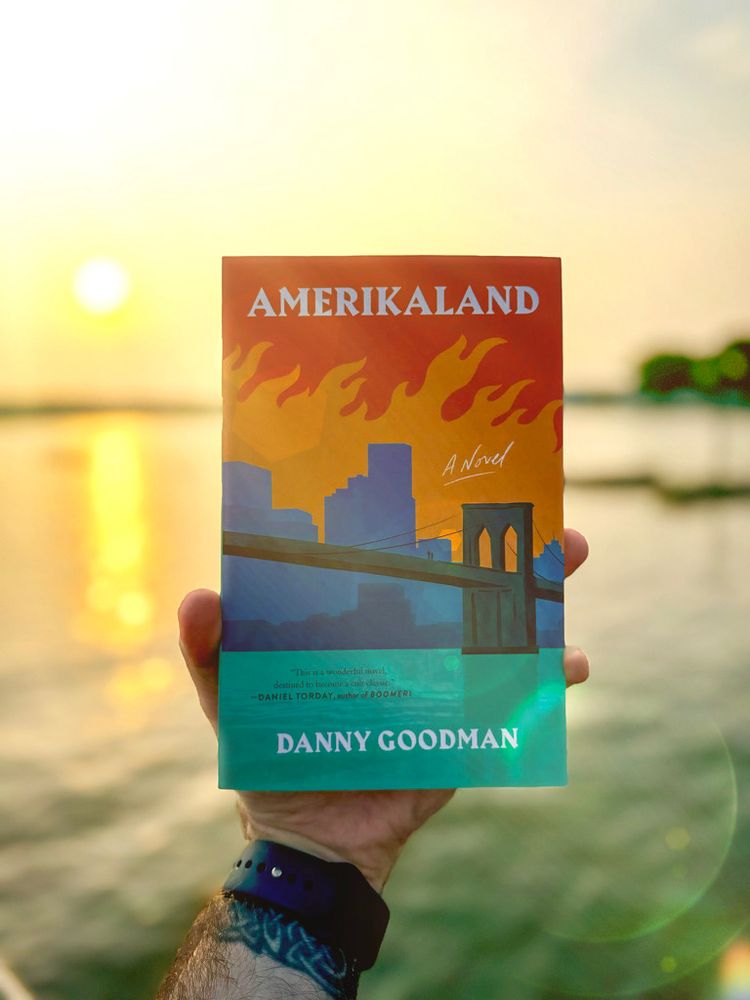 The image features the author holding a book titled "AMERIKALAND" in front of a serene body of water during sunset. The book cover has an illustration of vivid orange flames rising above a cityscape silhouette in blue, with a bridge in the foreground. The background showcases a soft, glowing sunset with a bright sun near the horizon, creating a warm and relaxing ambiance. The person holding the book is wearing a dark watch with a tattoo below it.