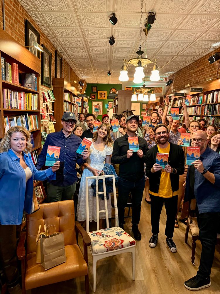 A happy group of people pose together in a bookstore, each holding up a copy of AMERIKALAND, surrounded by shelves filled with books.