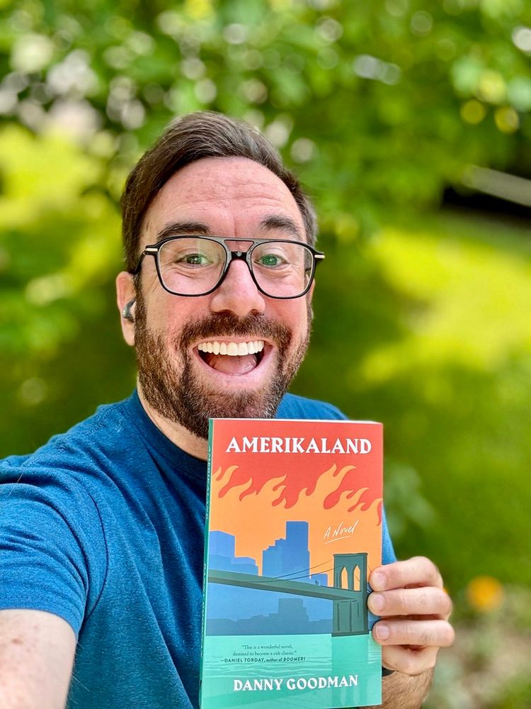 The image shows the author outdoors, smiling widely, and holding up a book titled "Amerikaland" by Danny Goodman. The individual is wearing black-rimmed glasses and a blue shirt. They have short dark hair and facial hair. The book cover features a stylized city skyline with flames above it, depicted in shades of blue, orange, and green against a red background. In the background, out-of-focus greenery suggests a garden or park-like setting with bright, diffused light.