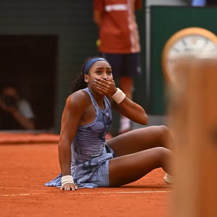 Coco Gauff sits on a clay court, covering her mouth with one hand, appearing emotional after winning the French Open.