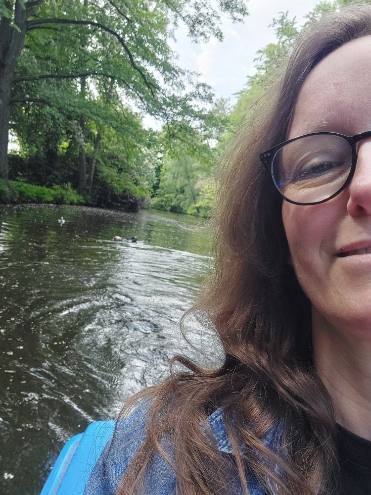 In der rechten Bildhälftegucke ich gut gelaunt ins Bild, befinde mich auf dem Wasser, am Rande eines Parks. Im Hintergrund schwimmt ein Wasservogel, eingerahmt ist das ganze von blühenden Bäumen und saftig grünem Blattgrün. 