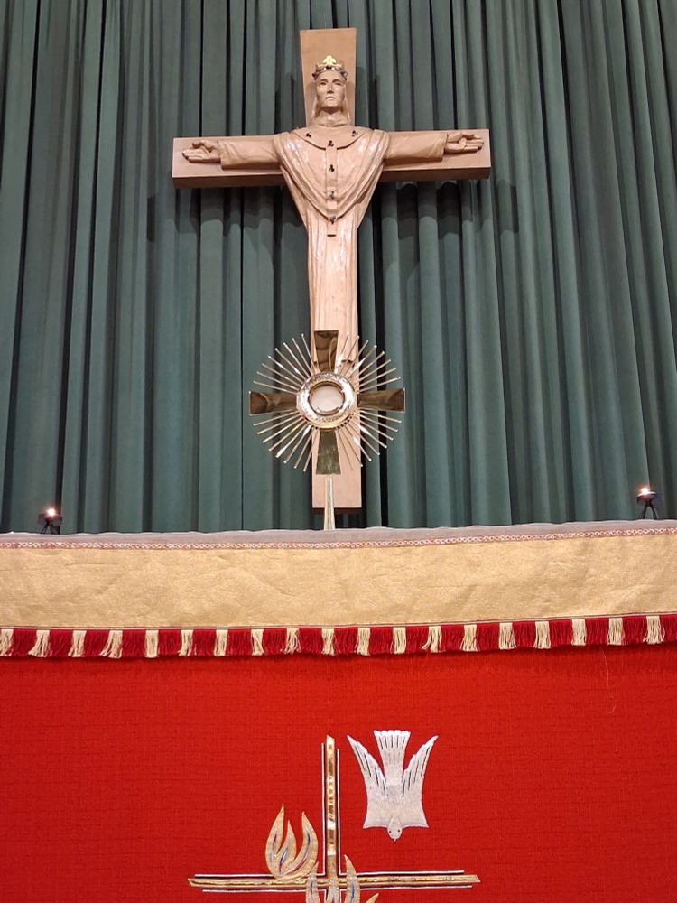 The blessed sacrament in a gold monstrance on an altar with a red altar frontal. Behind it is a large carved wooden Christus Rex in front of a dark green curtain. 
