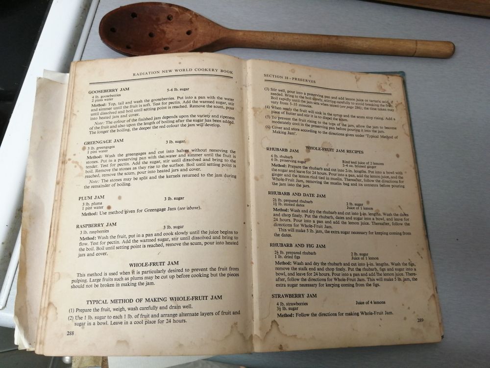 An old and very stained cookery book open at recipes for jam, with an old and stained wooden spoon next to it. 
