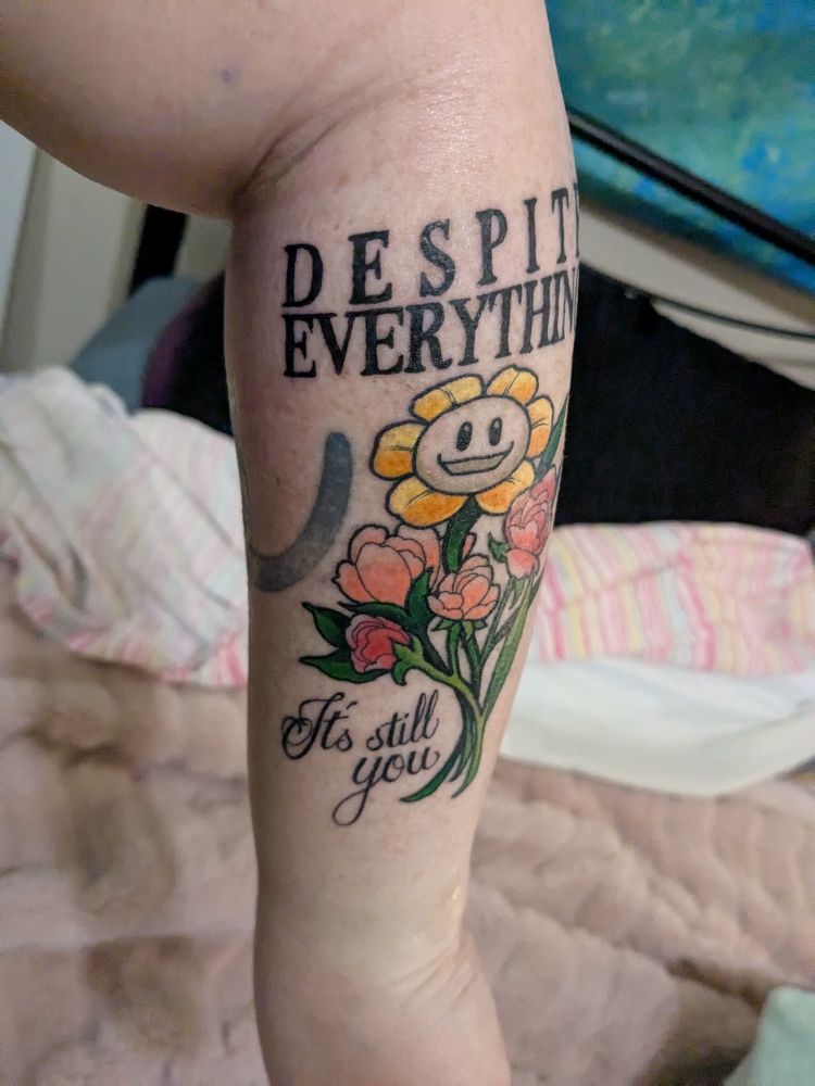 A fresh tattoo on a small bouquet of warmly coloured flowers featuring Flowey in the middle. Above says "Despite everything" and below says "it's still you"