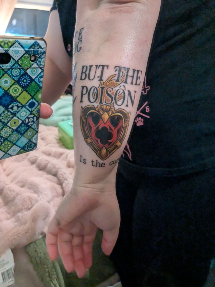 Fresh tattoo on the back of the forearm. A red sacred heart with church window architecture, framed with gold that says "But the poison" above and "is the cure" beneath.