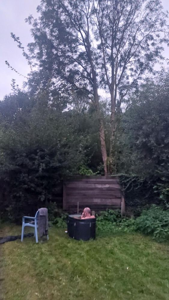 Op een gemaaid grasveld staat een blauw stoel met badjas. Daarnaast een zwart badje met een naakt persoon met kaal hoofd er in. Op de achtergrond struiken en bomen.