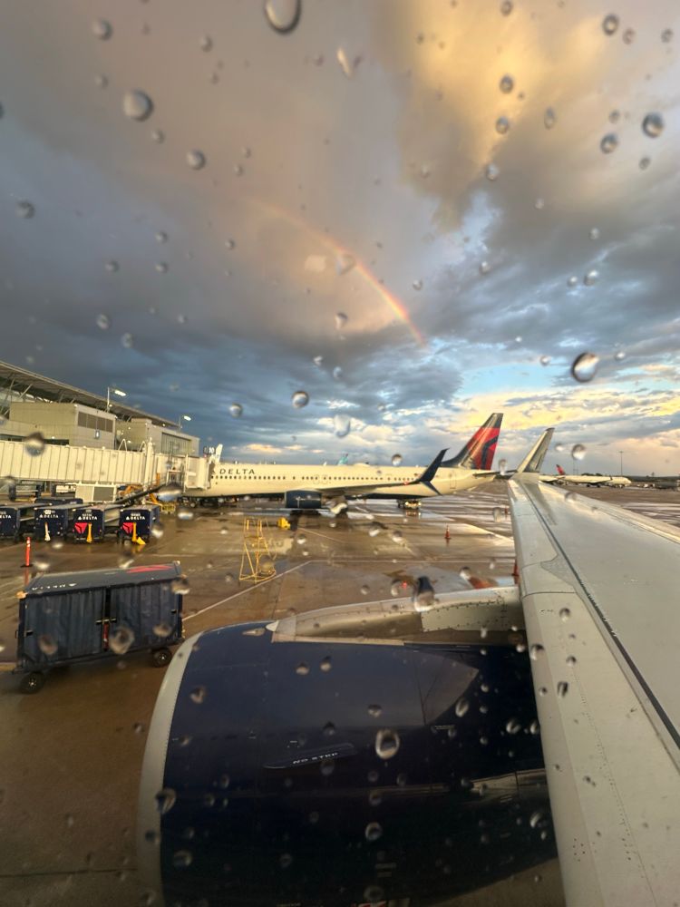 A view outside my airplane window, there are raindrops on the window but blue skies are starting to peak through the gray rain clouds and a rainbow can be seen in the sky. The ground is wet below a Delta plane across my plane
