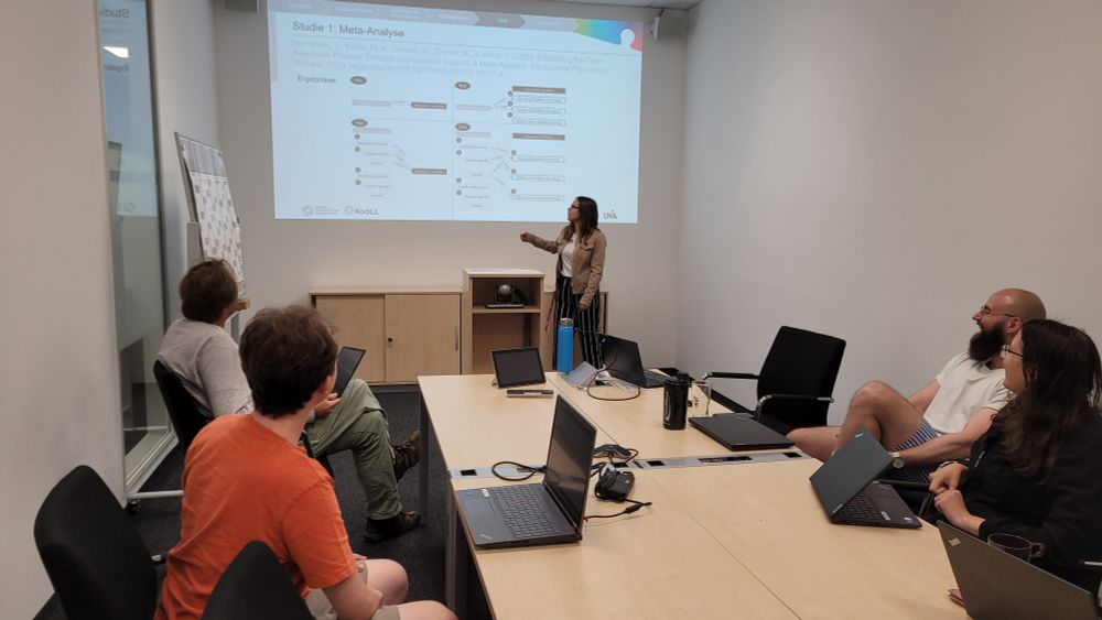 Das Foto zeigt Julia Hornstein, wie sie Forschungsergebnisse zu Peer Feedback im Team-Meeting des DigiLLabs der Uni Augsburg präsentiert. Sie steht vor einer Präsentationsfolie mit einer wissenschaftlichen Grafik, die die Ergebnisse der Meta-Analyse zusammenfasst.