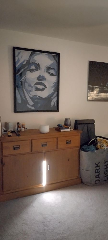A piece of art on a wall above a wooden dresser. The art is a woman's face constructed from pieces of denim. To the right is part of another art work. Between them on the floor is a laundry hamper in front of a trouser press.