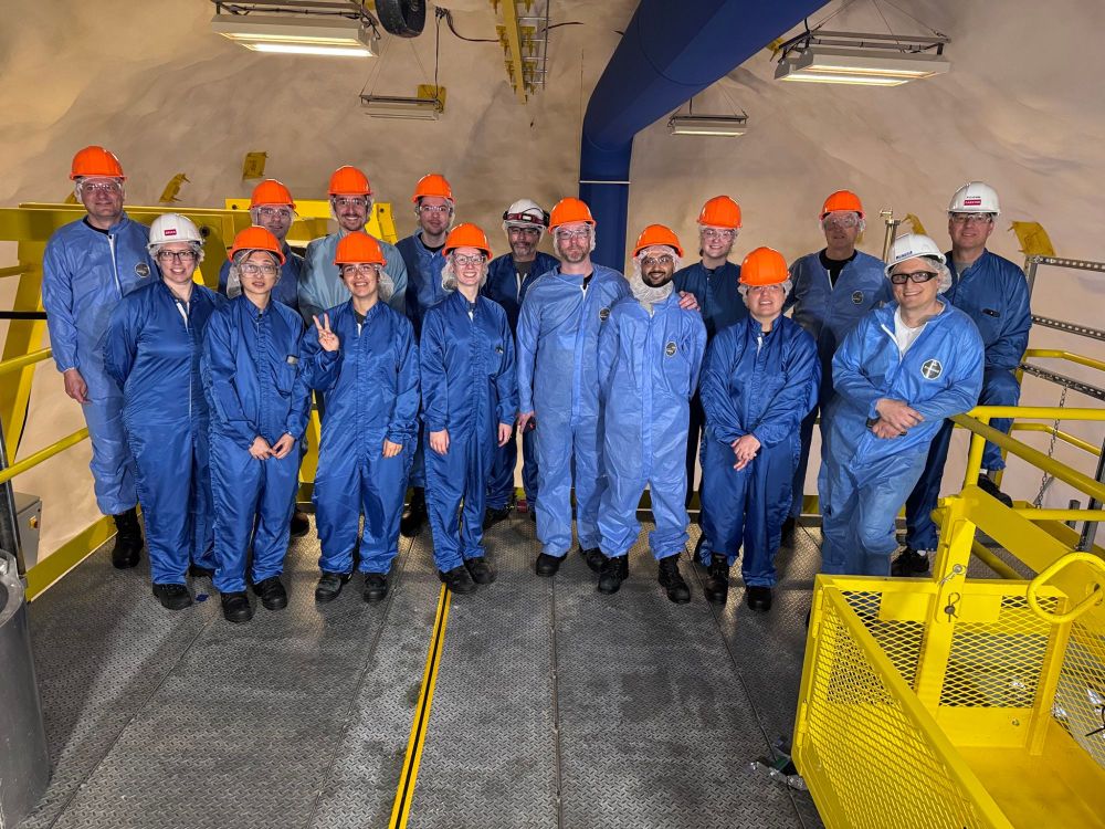 Sixteen scientists in blue clean-room suits, safety glasses and orange or white hard hats pose shoulder-to-shoulder on a yellow metal platform inside an underground cavern, beaming at the camera.