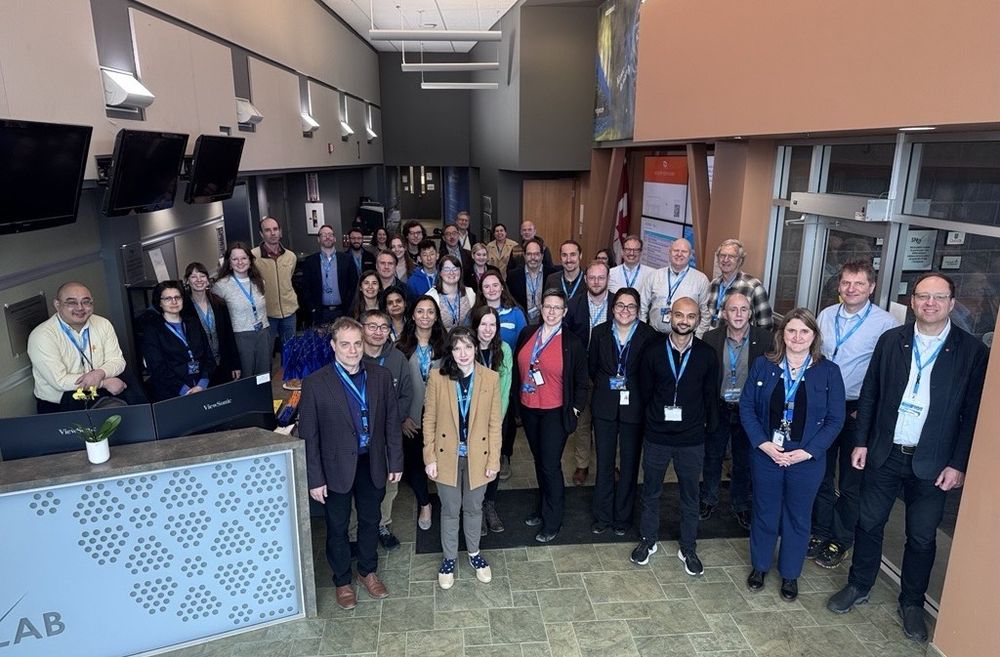 Roughly forty workshop participants wearing blue lanyards cluster in the atrium of a modern research facility, smiling up at the camera; a reception desk with monitors is on the left and glass entrance doors on the right.