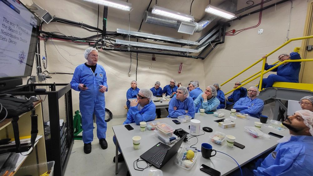 In a clean-room, a speaker in blue coveralls and a hairnet stands beside a slide titled “The way forward in the search for neutrinoless double beta decay” while about a dozen similarly suited colleagues sit at tables with boxed lunches, notebooks and laptops, listening intently.