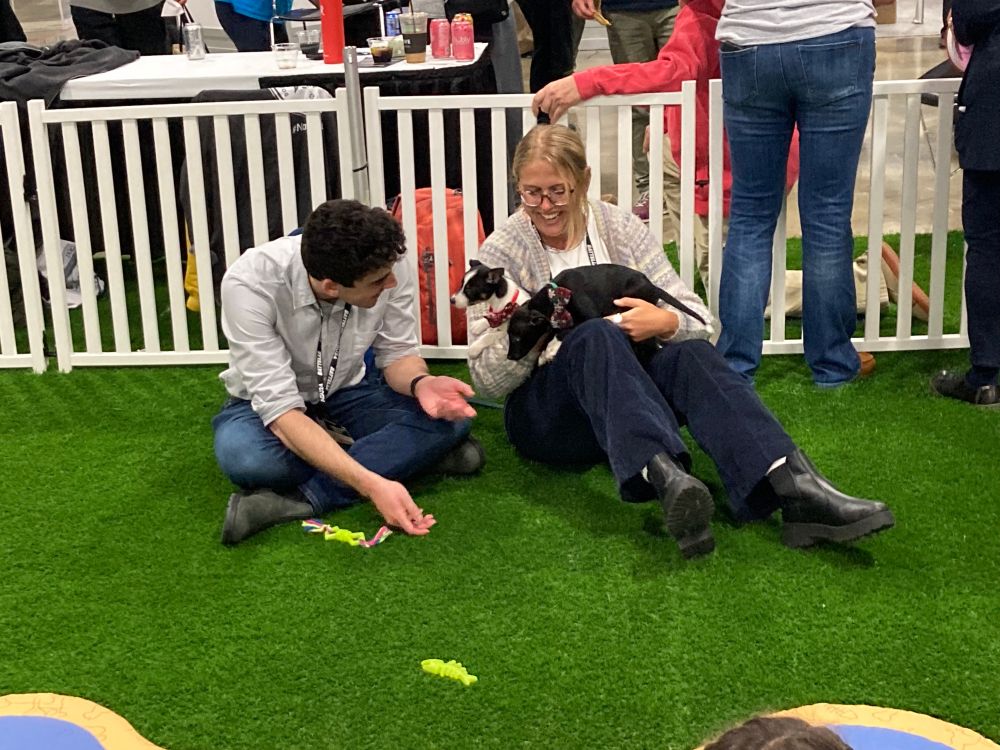Two guys sitting on fake grass surrounded by a low white picket fence; one guy has two puppies in his lap and both men are looking joyfully at the little pooches. 