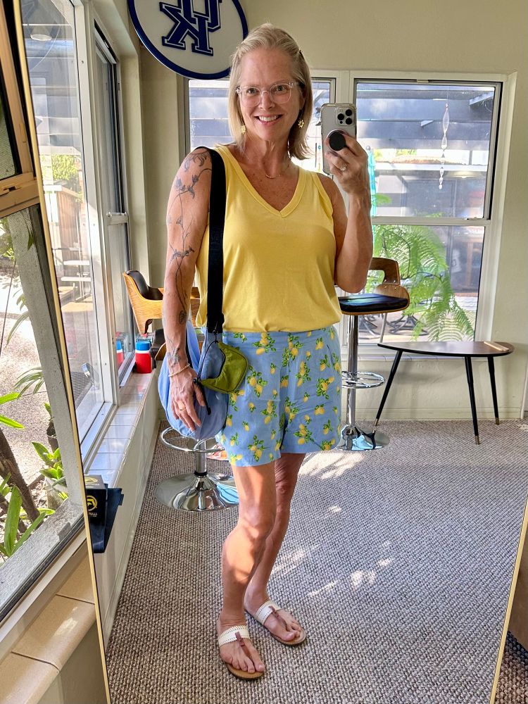 A mirror selfie wearing clear acrylic glasses, a yellow tank top, blue shorts with little lemons all over them, and white and leather sandals. I am also wearing a blue Baggu bag. 