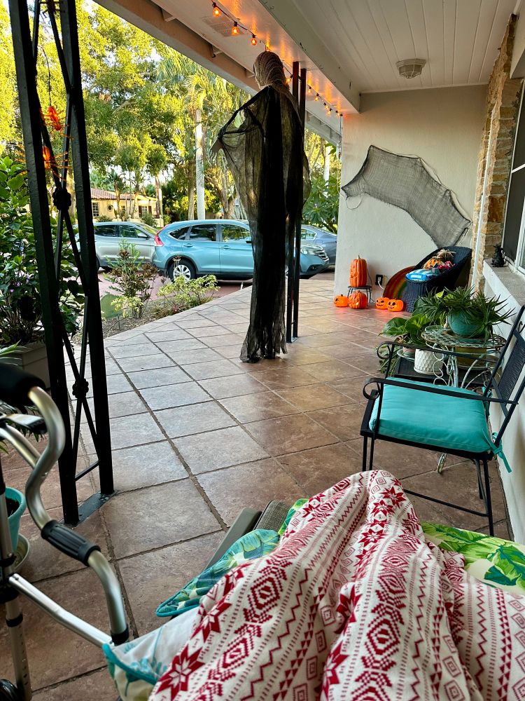 Me sitting on the front porch with my legs covered by a blanket and my walker next to me. Halloween decorations can be seen on the porch as well as plants and cars parked in the driveway. 