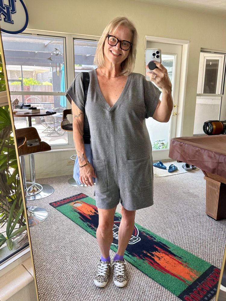 A mirror selfie wearing a gray knit shorts romper, tortoise shell horn rimmed glasses, purple socks and leopard print sneakers. Carrying a blue purse. 