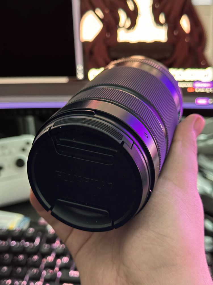 Me holding a large telephoto zoom lense with the cap on that says “Fujifilm”, with the background heavily in blur, keyboard, controller and a stream open vaguely in the background 