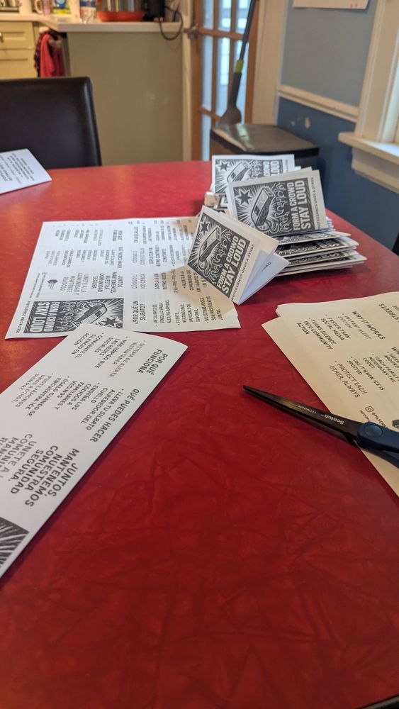 A red table is covered with numerous printed zines, in Spanish , about using whistles to warn about ICE and a pair of scissors