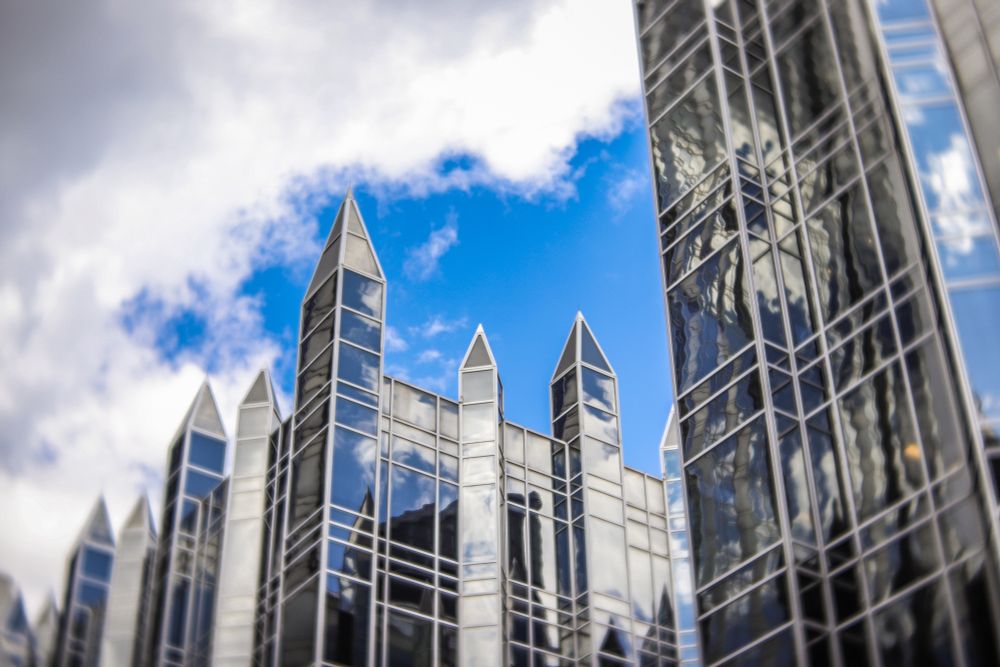 Glass skyscrapers with pointed, castle-like spires reflect the sky and clouds, set against a bright blue sky with patches of white clouds.