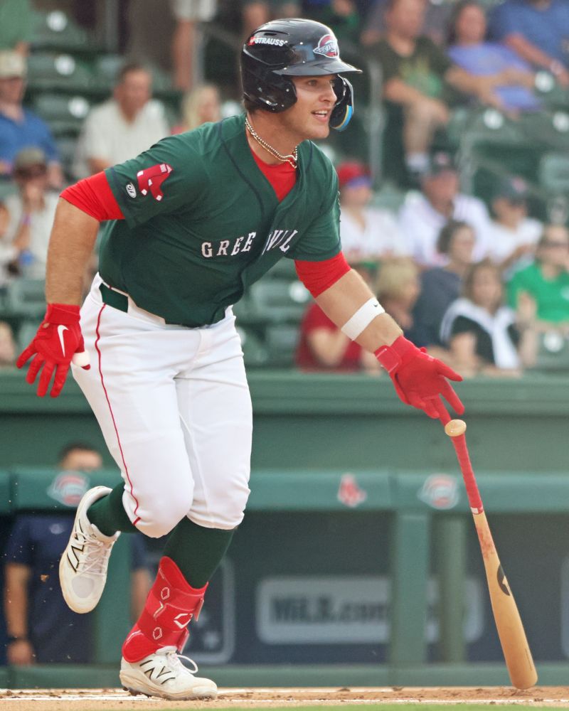 Henry Godbout, Red Sox 2025 draftee and newly minted Greenville Drive second baseman, drops his bat as he starts the run to first base after hitting a single. He wears a green jersey with GREENVILLE across the front in white, white pants with his green socks, and red batting gloves and ankle guard, with a red shirt showing at the sleeves and neckline, and white New Balance cleats. His face has an expression of boyish enthusiasm.