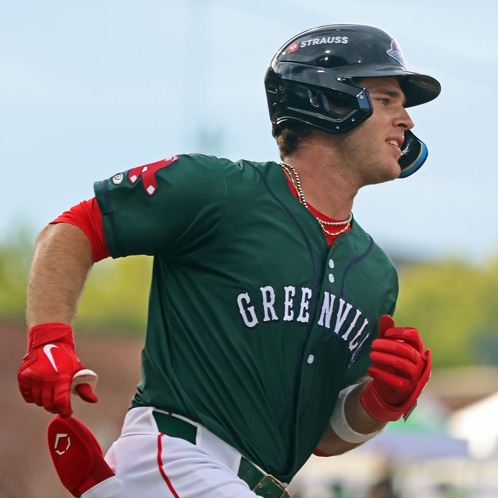Henry Godbout is shown from the waist up on his way to first base after hitting a single. He wears a green jersey with GREENVILLE across the front in white, white pants, and red batting gloves, with a red shirt showing at the sleeves and neckline, and a doubled silver chain. His face has an expression of confidence.