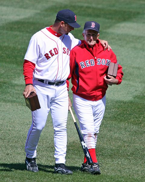 Red Sox pitcher Tim Wakefield (in his home white Red Sox uniform) walks across the Fenway grass with his 2007 World Series ring box in his right hand. HIs left arm is around the shoulders of Red Sox veteran Johnny Pesky, wearing a red Red Sox jacket over white pants, with a bat in his right hand and his own ring box tucked in his left arm.