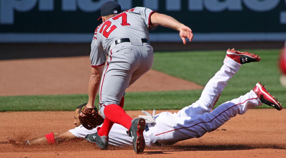 Mookie Betts, wearing the Red Sox home white uniform, slides on his torso, legs still in the air and dirt flying as he steals third base after a successful steal of second; he is helmet-less and his expression is determined. Nationals first baseman Ryan Zimmerman races away from the camera toward Mookie with glove down hoping to make the tag in time. (He didn't.) The base of Fenway's iconic Green Monster is visible in the background.