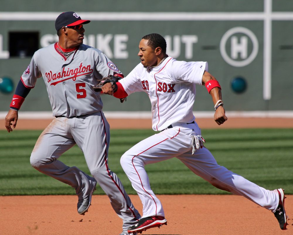 Mookie Betts, wearing his Red Sox home white uniform, races to third base after a successful steal of second; he is helmet-less and his expression is determined. His stride is fully extended. Washington Nationals third baseman Yunel Escobar runs with him looking back to second base for a possible throw, but his resigned expression tells us that he won't get a throw in time to get Mookie out. Fenway's iconic Green Monster is visible in the background.