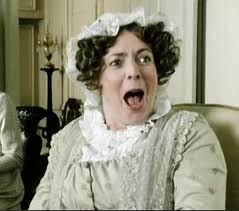 Mrs Bennet from the 1995 BBC Pride and Prejudice series. She has her mouth wide open in humorous alarm, and a strange gleam in her eyes. She is wearing a white lace cap and collar with a pale green, slightly shiny dress, and is seated in a drawing room.