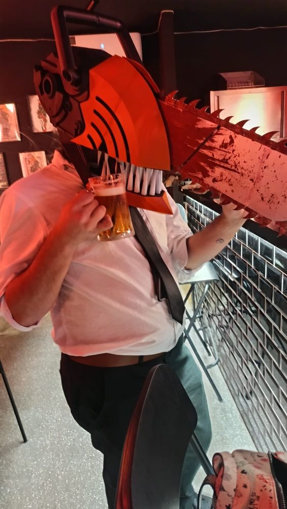 Mesma roupa e capacete de antes, mas em um bar, na frente do balcão, com uma cerveja na mão e um canudo levando da cerveja pra dentro da boca, entre os dentes.
