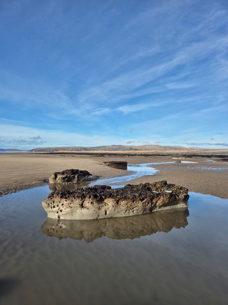 View of a sandy beach with sand dunes and hills in the background. Sky is blue with some cloud. In the foreground, in a shallow tidal pool there is an exposure of pale grey clay with a thin covering of dark brown peat on top.