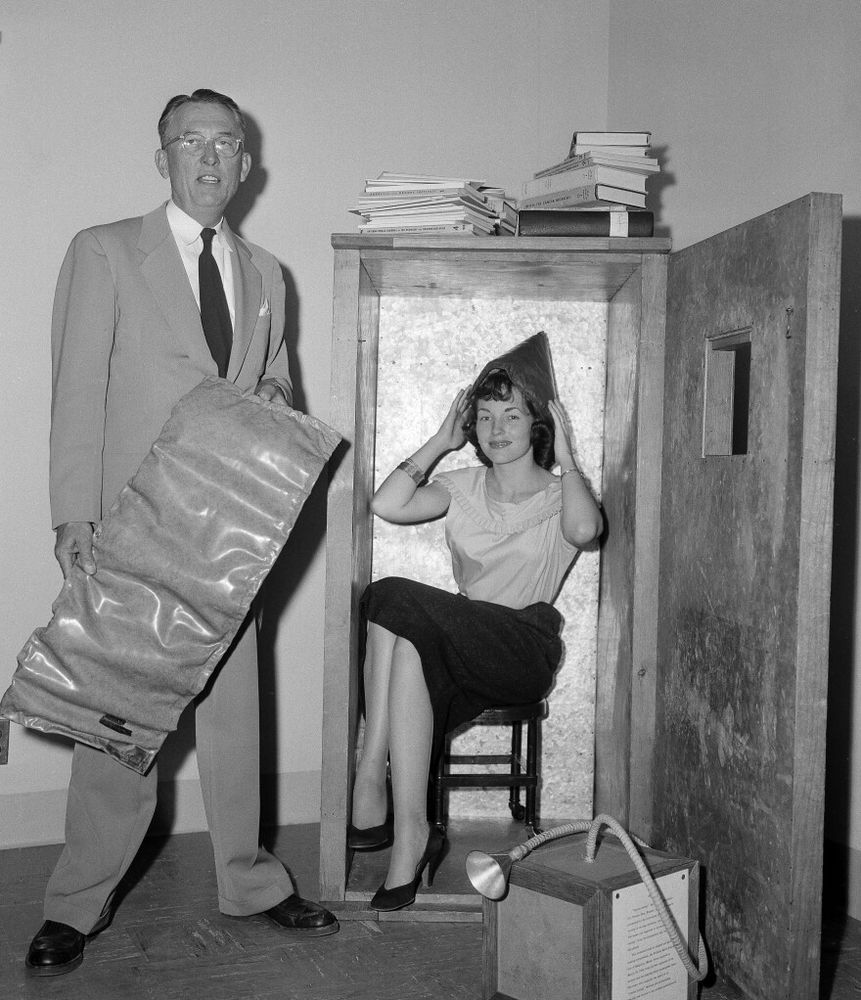 A black and white photo of Wilhelm Reich standing next to a woman sitting inside of one of his quack "Orgone accumulator" boxes from the 1950s. It is a small wooden box, lined with metal and with a stool inside. The door is open and has a small window.