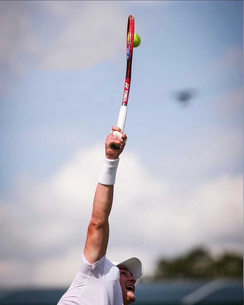 Foto em close do João Fonseca sacando, mostrando a deformação da raquete e da bolinha de tênis no momento do contato.