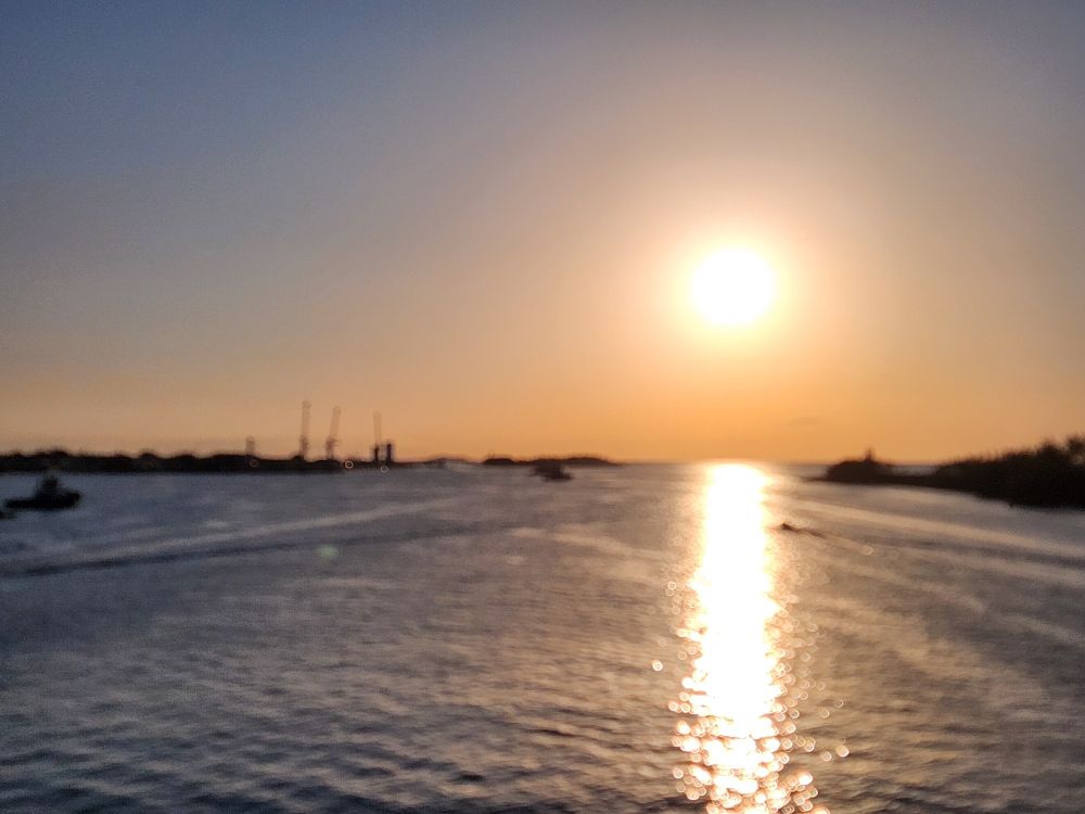 "Blurry view of the sun setting over a calm body of water, with silhouetted landmasses and cranes on the horizon, and the golden light reflecting across the surface."