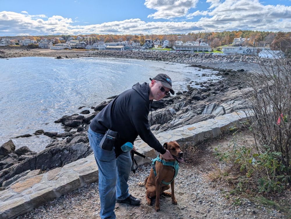 A photo of a white man in jeans and a baseball cap with a brown dog standing on the coast of an inlet with a small town behind them. 