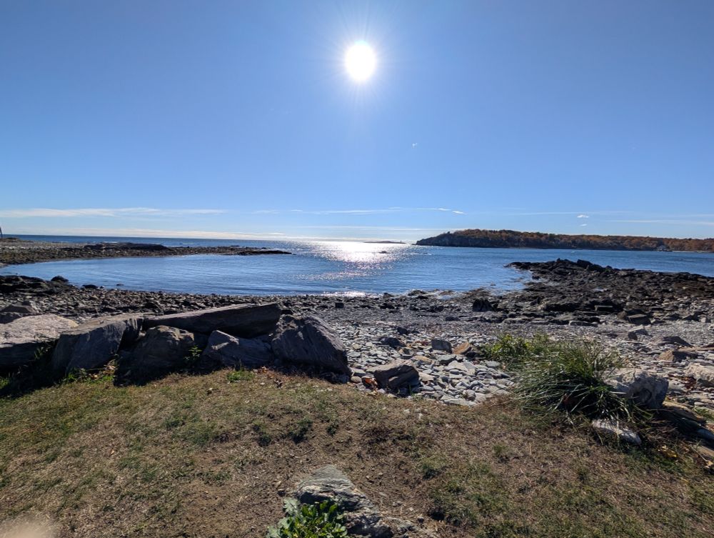 A photo of a rocky Cove of water with a very blue sky and a bright syn in the center of the sky. 