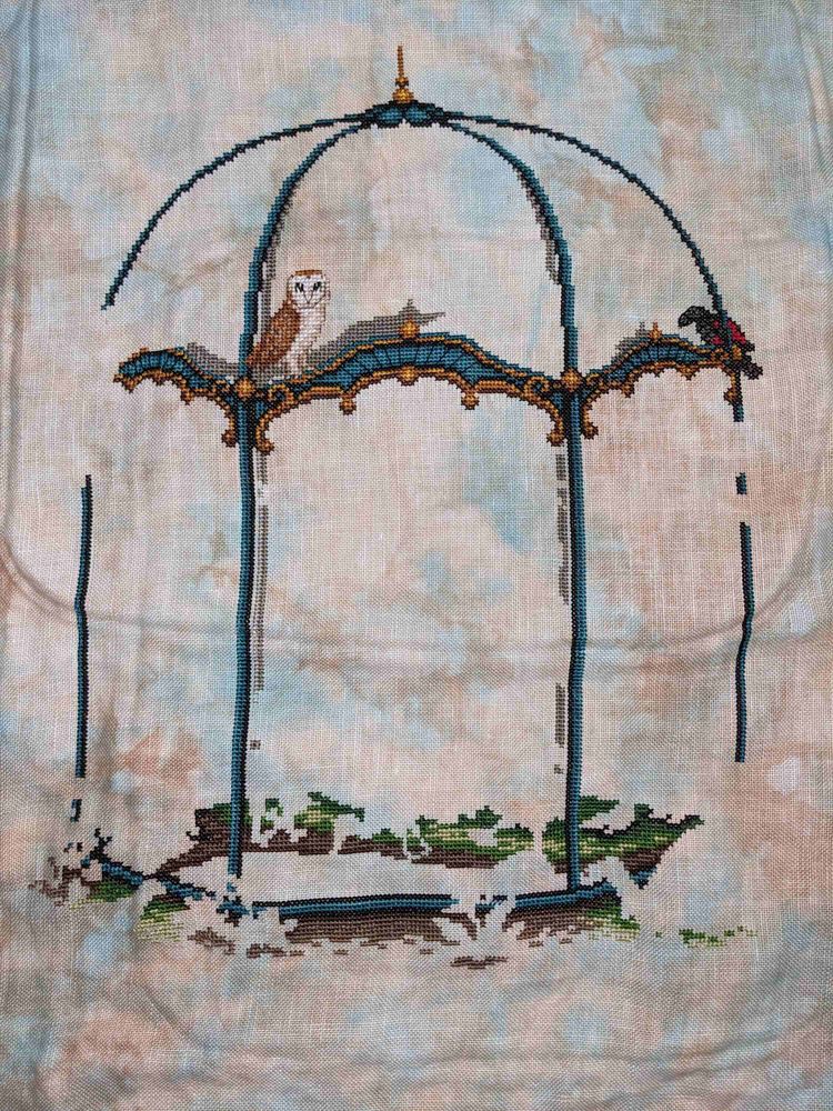 The first part of Extraordinary Birds SAL finished - showing an aviary with an owl perched near the top and a black and red parrot perched off to the side on blue and tan linen.