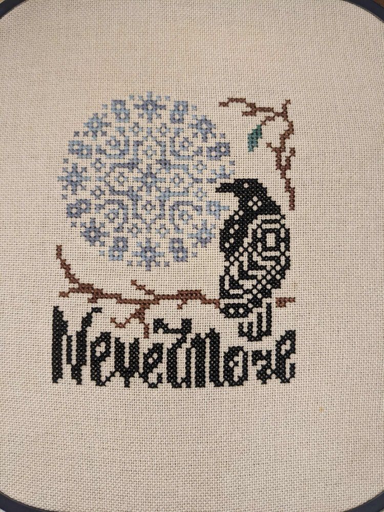 A finished cross stich of a raven sitting on a brance with a blue moon in the to left corner. Nevermore is written below the branch.