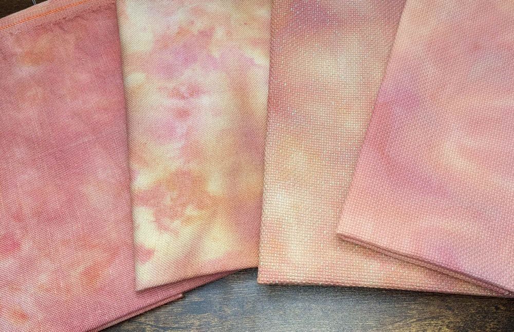 Four pieces of fabric in a sunset palette of pinks, oranges, and yellows. Shown are linen, evenweave, opalescent Aida, and Aida.