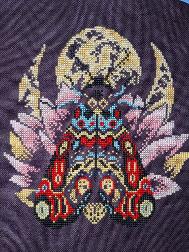 A partially completed cross stitch on deep purple evenweave. The design is a luna moth with wings down on top of a pink petalled flower with a moon in the top background.