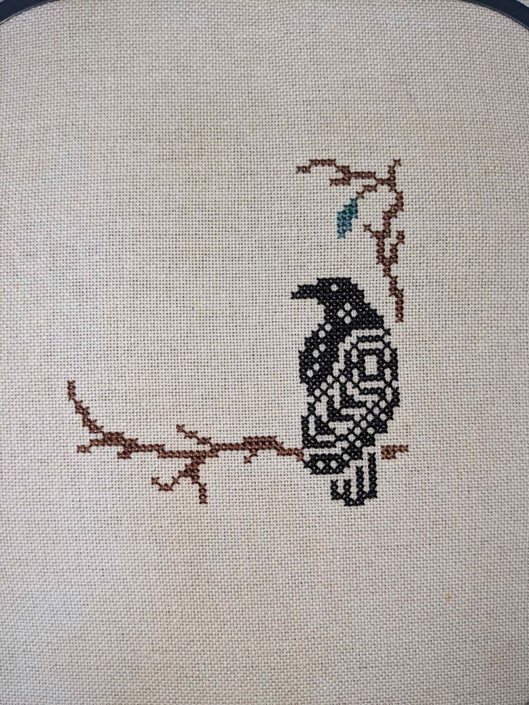 A small cross stitch showing a raven perched on a branch.
