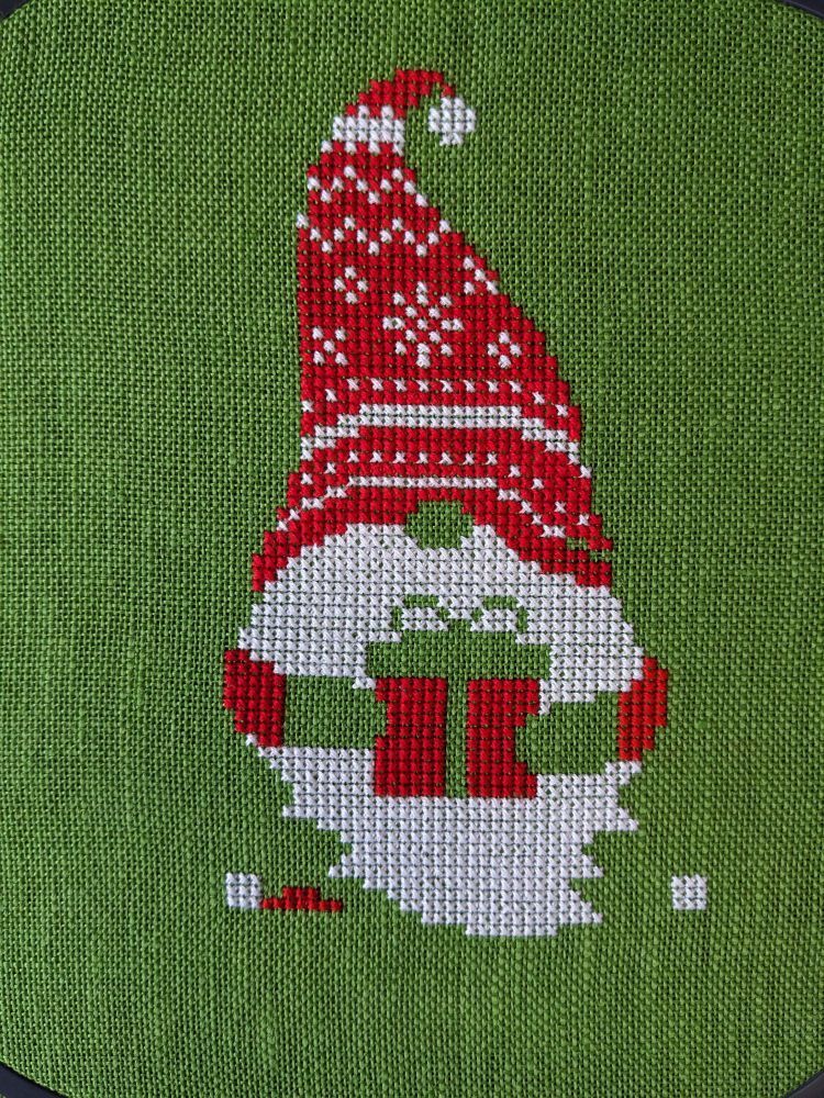 A Christmas gnome with a tall red stocking cap with white details, a long white beard covering pretty much all of him, holding a red present. Design is on green linen fabric.