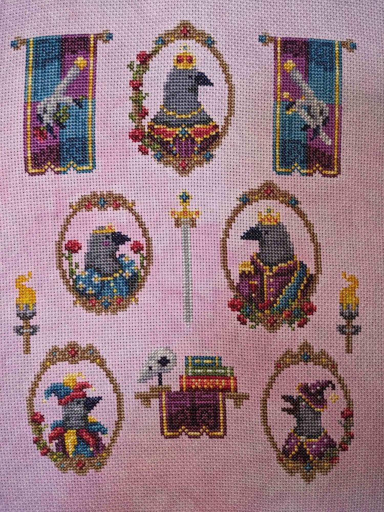 A completed cross stitch piece on dusty pink fabric showing a royal crow court in frames, two pennants, and a sword.