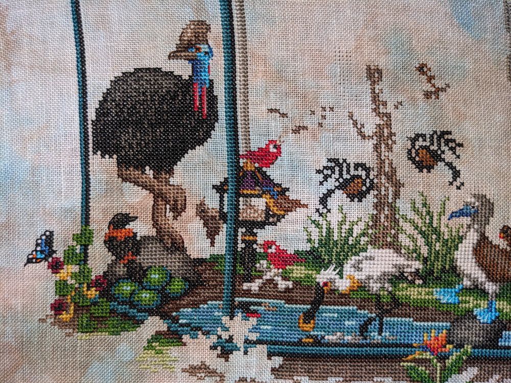 Portion of a cross stitch piece showing a variety of birds and plants in a giant aviary.