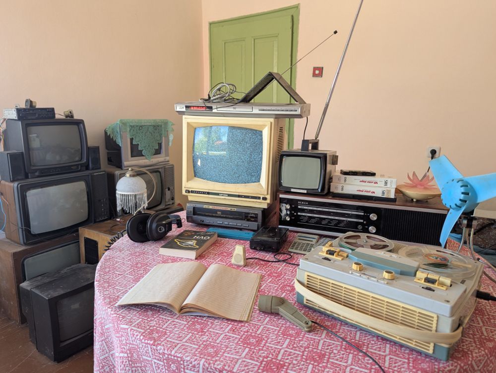 pic of a table with a reel to reel recorder, notebook, two microphones, dvd player, two tv sets, radio, a fan and some other thingies.
back left wall is full of tv sets stacked on top of each other
