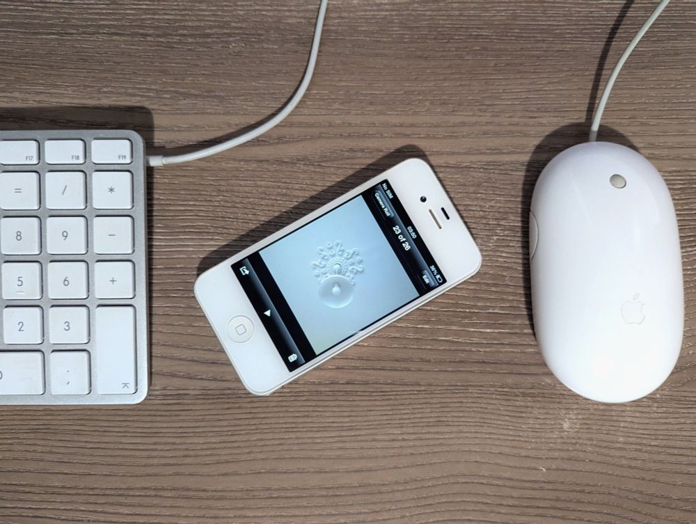 Picture of a white iPhone 4S running iOS 6. With an image open of a white lamp on a white ceiling. There's an A1243 keyboard on the left of it and a Mighty Mouse on the right 