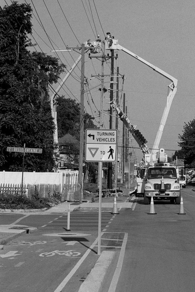 A black-and-white photo of two cherry pickers blocking a road and bike lane to work on a utility pole.