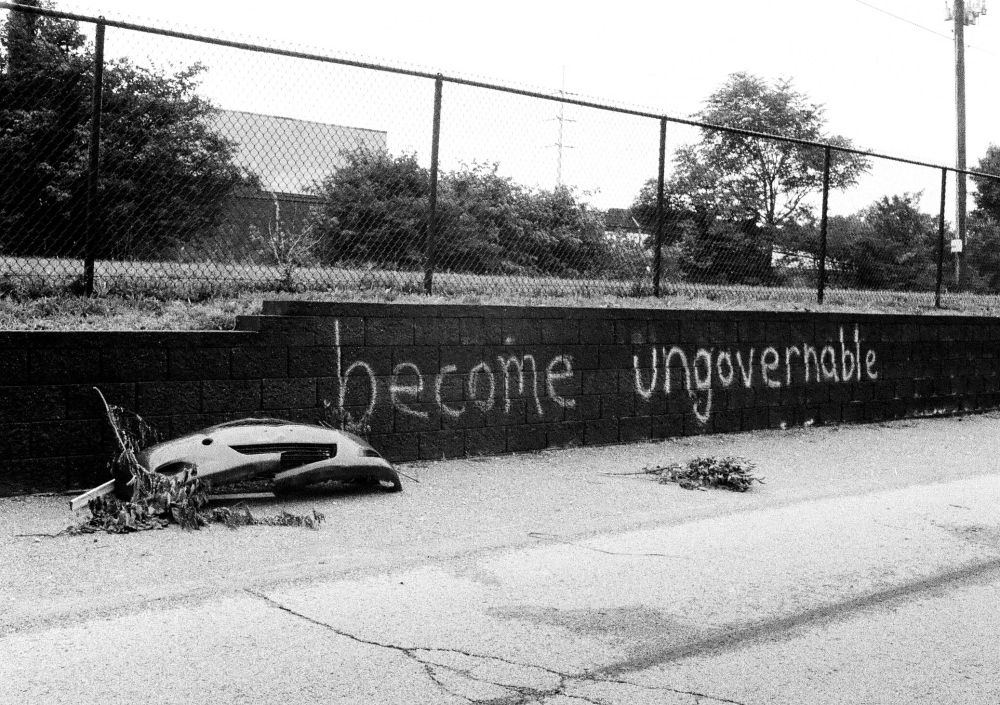 A black-and-white photo of "become ungovernable" graffitied in large letters on a retaining wall next to a wrecked car bumper that is nowhere near a road.