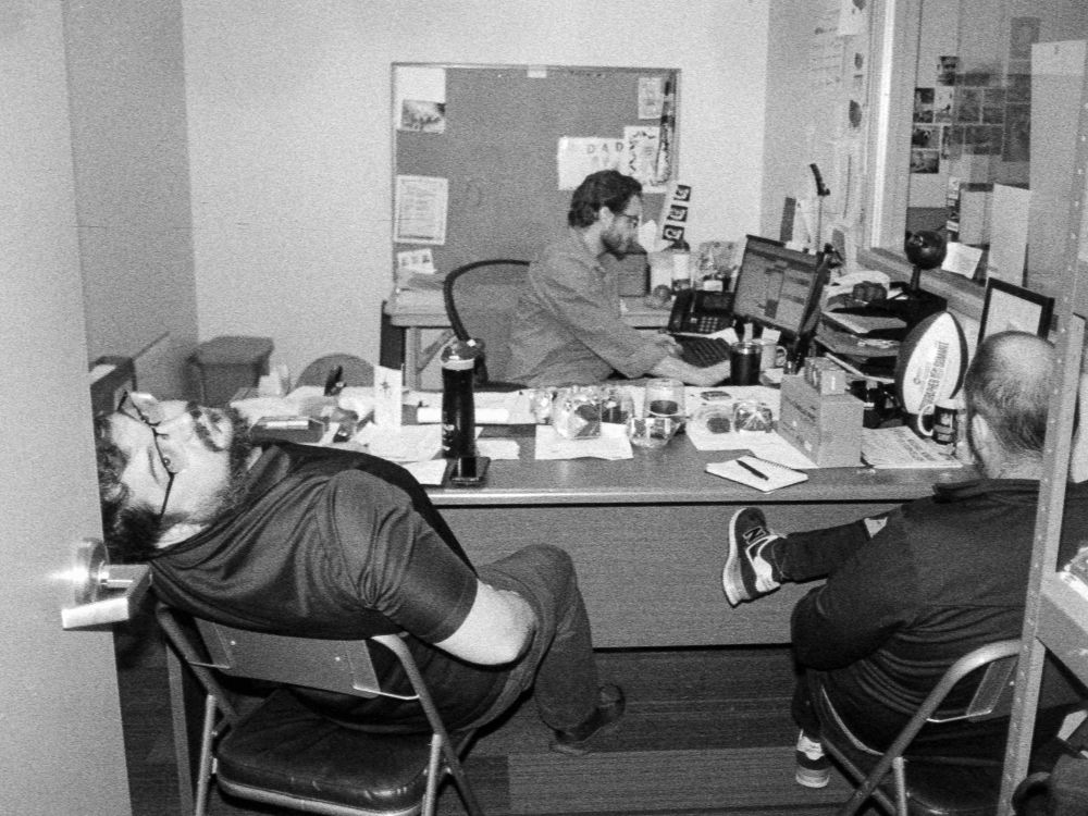 A black-and-white flash photo of two men on one side of a desk across from a third in a small office. One man is leaning back in a chair to look up at the photographer.