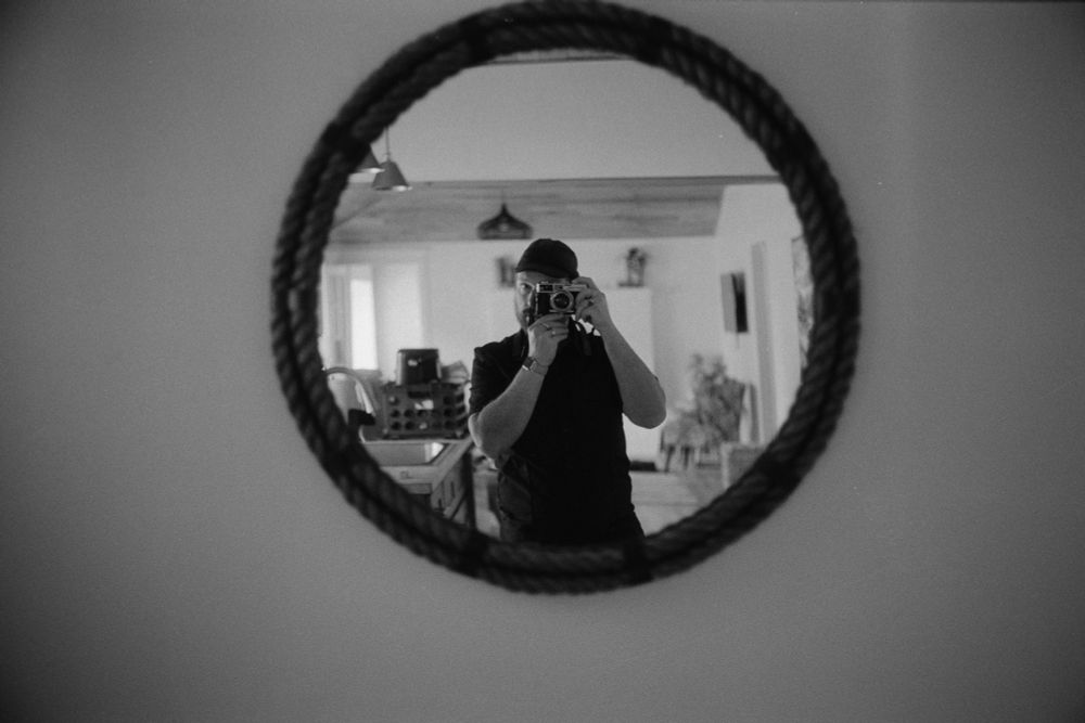 A black-and-white self-portrait of a male photographer using a vintage rangefinder reflected in a circular mirror wrapped with two rings of fat rope.
